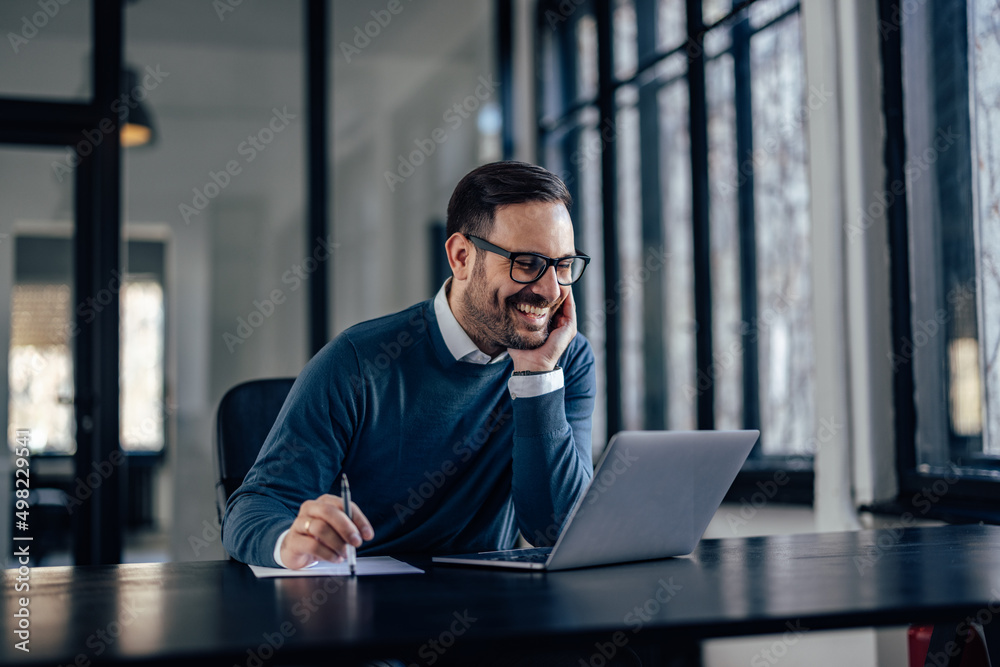 Smiling man with glasses on, using a laptop, at his office. Smiling man with glasses on, using a laptop, at his office.