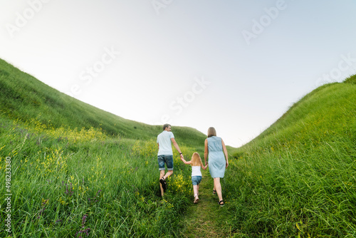 Mom, dad, and daughter walk back in the green grass. Happy young family spending time together, running on nature, on vacation, outdoors. The concept of a family holiday. Back view.