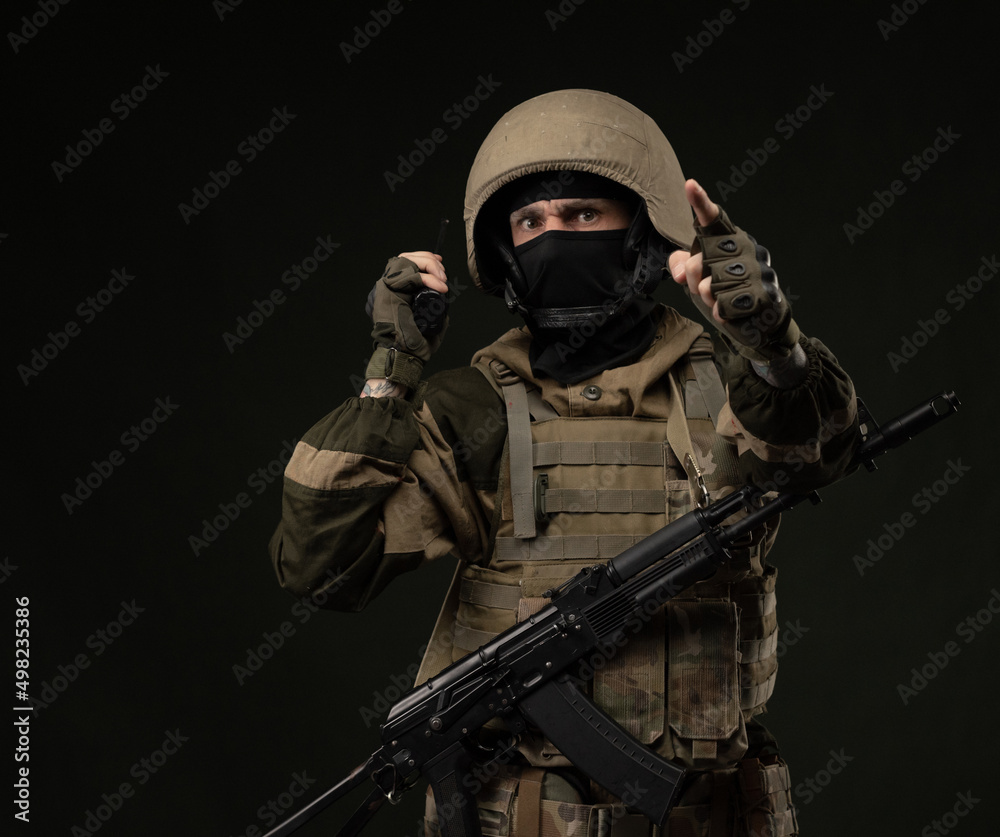 a Russian soldier in military clothes with a Kalashnikov assault rifle on a dark background takes orders on a walkie-talkie a Russian soldier in military clothes with a Kalashnikov assault rifle on a dark background takes orders on a walkie-talkie