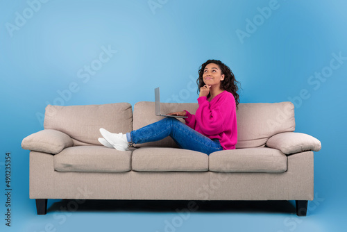 Thoughtful young black woman in casual wear working online, sitting on couch and using laptop on blue studio background