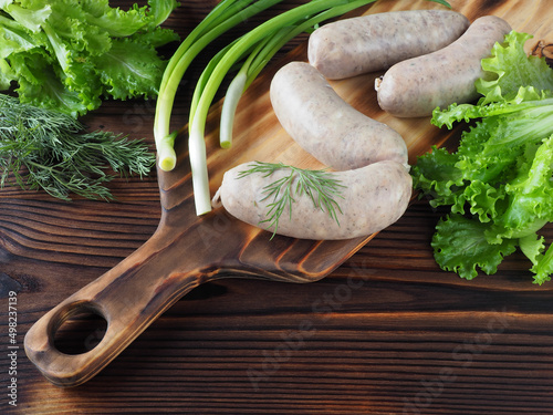 Fotobehang Raw bavarian sausages on the kitchen board, fresh greens on a dark wooden table, closeup, flat layout, copy space