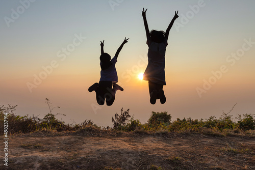 Silhouette of happy two children jumping on top mountain sunset background.