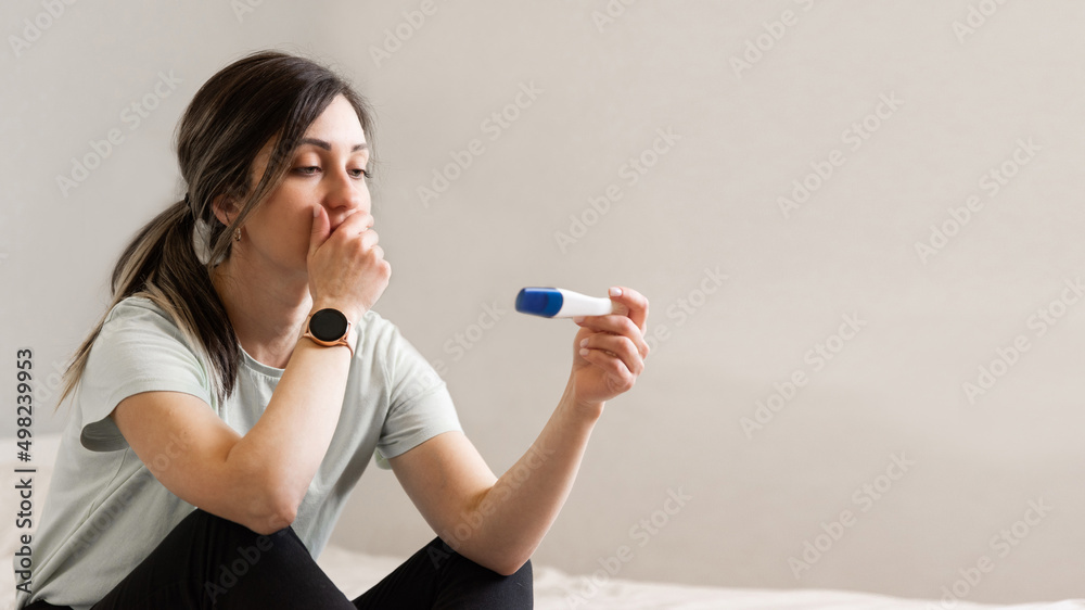 the woman found out about an unwanted pregnancy. A woman with a pregnancy test in her hands is sitting on the bed in her apartment