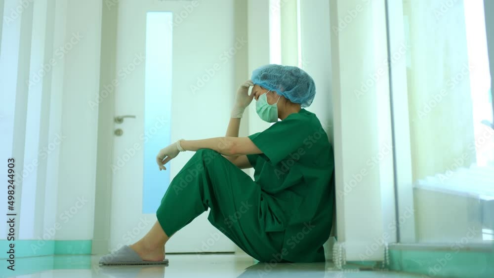 Side view of stressed and overworked female doctor medical worker surgeon sitting on the floor near the operating room in hospital and take off the mask Side view of stressed and overworked female doctor medical worker surgeon sitting on the floor near the operating room in hospital and take off the mask