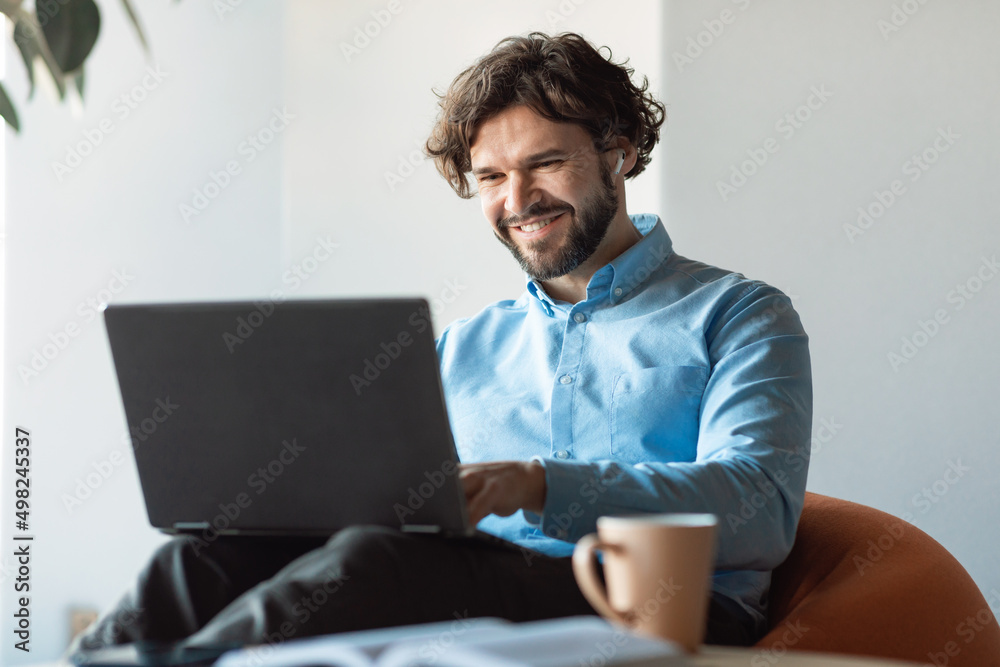 Smiling businessman using laptop sitting on beanbag in office Smiling businessman using laptop sitting on beanbag in office