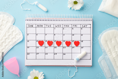 Top view photo of red heart marks on the calendar hygienic pads tampons pink menstrual cup and camomile buds on isolated pastel blue background photo
