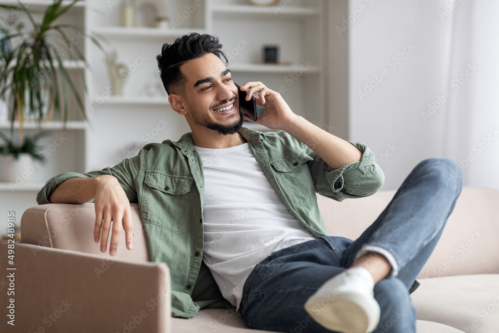 Cheerful arab guy having phone call while relaxing on couch at home
