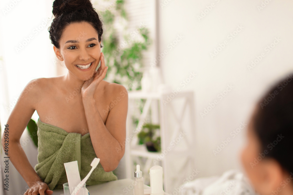 Beautiful young woman in towel looking at herself in mirror, touching her face and smiling at home, free space Beautiful young woman in towel looking at herself in mirror, touching her face and smiling at home, free space