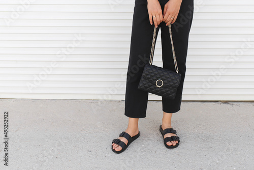 Bag in women's hands. Woman wearing black pants, bag with chain and flat sandals standing outdoor. Details of stylish trendy basic minimalistic casual outfit. Street fashion. Women's legs, no face.