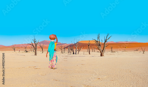 Woman in traditional green dress carrying heavy jug of water on her head and walking on the Dead trees in Dead Vlei - Sossusvlei, Namib desert, Namibia