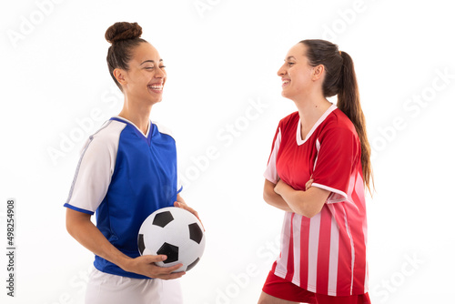 Smiling caucasian and biracial young female soccer players talking standing against white background