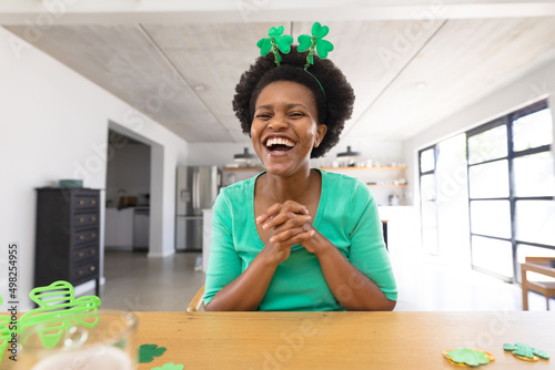 Happy african american mid adult woman wearing shamrock headband sitting with hands clasped at home