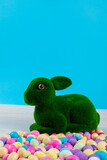 Artificial moss bunny with colorful candy easter eggs on table against blue background, copy space