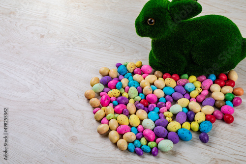 Artificial moss bunny with colorful candy easter eggs on table with empty space