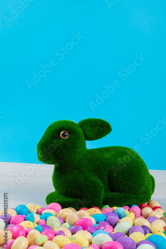 Artificial moss bunny with colorful candy easter eggs on table against blue background, copy space