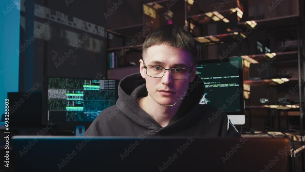 A young guy with glasses is working on a laptop. Behind him are 2 monitors with changing digital codes. This is a programmer, he gets upset because of failure A young guy with glasses is working on a laptop. Behind him are 2 monitors with changing digital codes. This is a programmer, he gets upset because of failure