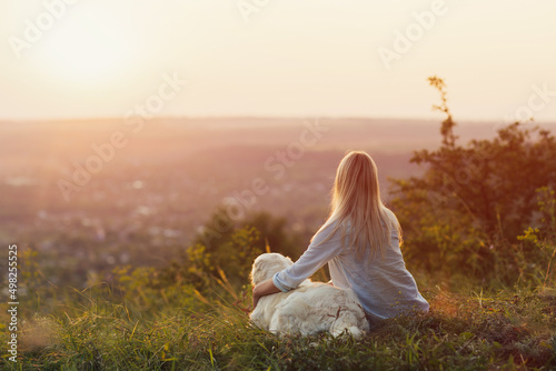 Blonde woman hugging dog while sitting on the hill at sunset. Beautiful golden retriever and girl relaxing together. Lifestyle.