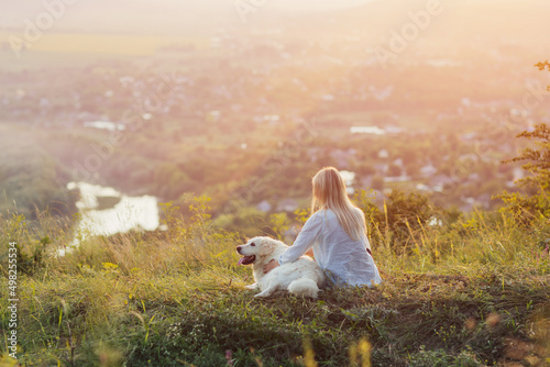 A young woman and a dog sitting on the hill and enjoying countryside landscape on a summer evening. A lady and her faithful friend.