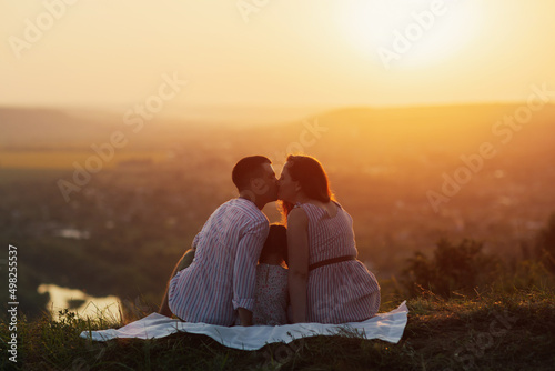 Husband kisses his wife while they sitting on the hill at sunset. Little daughter sits between the parents. Happy young family is resting in the countryside. a Bright sunny sunset.