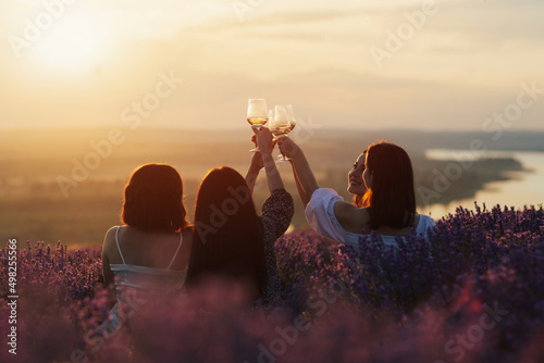 Carefree happy women enjoy moment together  meeting the sunset  spend weekends in the lavender field. They clinking glasses with wine.