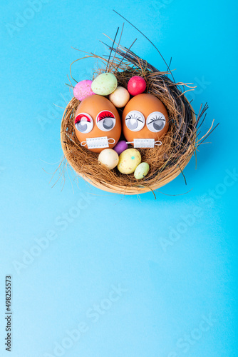 Easter eggs with mask and doodle eyes with candies and straws in nest on blue background