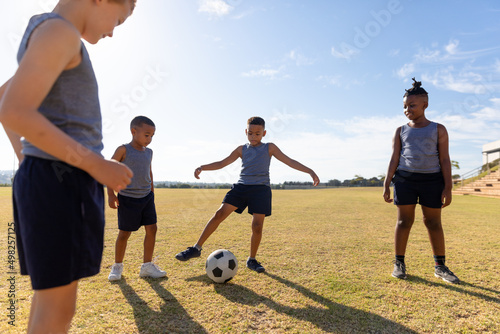 Multiracial elementary schoolboys looking biracial boy kicking soccer ball on field while playing