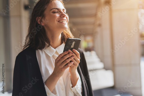 Business woman having good news from partners wia smartphone while walking near office building, Smiling girl chatting with friends while coffee break outdoor