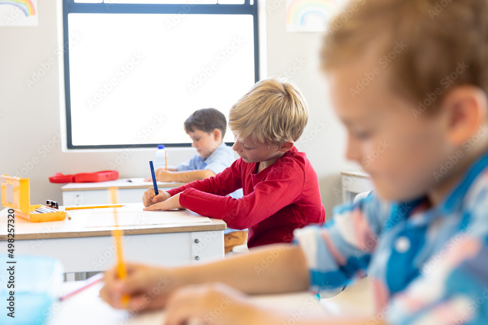 Multiracial elementary schoolboys writing on books while sitting at desk in classroom Multiracial elementary schoolboys writing on books while sitting at desk in classroom