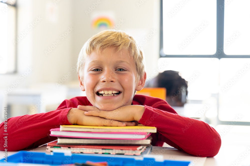 Portrait of smiling caucasian elementary schoolboy leaning on stacked books at desk in classroom Portrait of smiling caucasian elementary schoolboy leaning on stacked books at desk in classroom