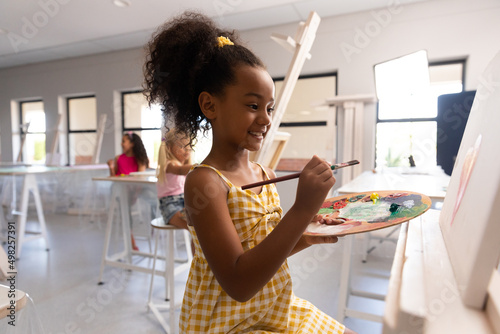 Smiling biracial elementary schoolgirl with palette and paintbrush painting on easel in school