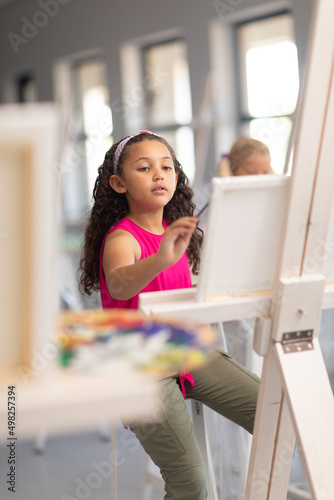 Cute biracial elementary schoolgirl painting on easel during drawing class in school