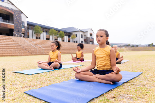 Multiracial elementary schoolgirls meditating while exercising on exercise mat at school ground