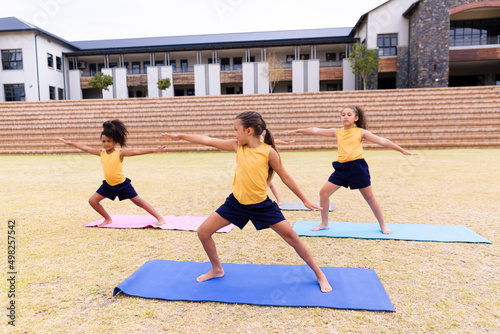 Multiracial elementary schoolgirls doing warrior 2 pose while exercising on yoga mat on ground