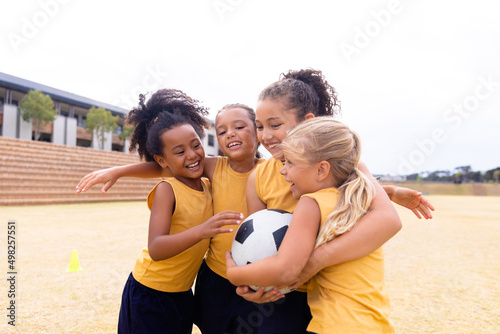 Cheerful multiracial elementary schoolgirls with soccer ball embracing while standing on ground