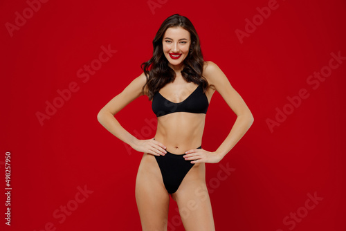 Young smiling sexy brunette woman 20s with perfect fit body wear black underwear stand akimbo arms on waist lok camera isolated on plain red background studio portrait. People female beauty concept