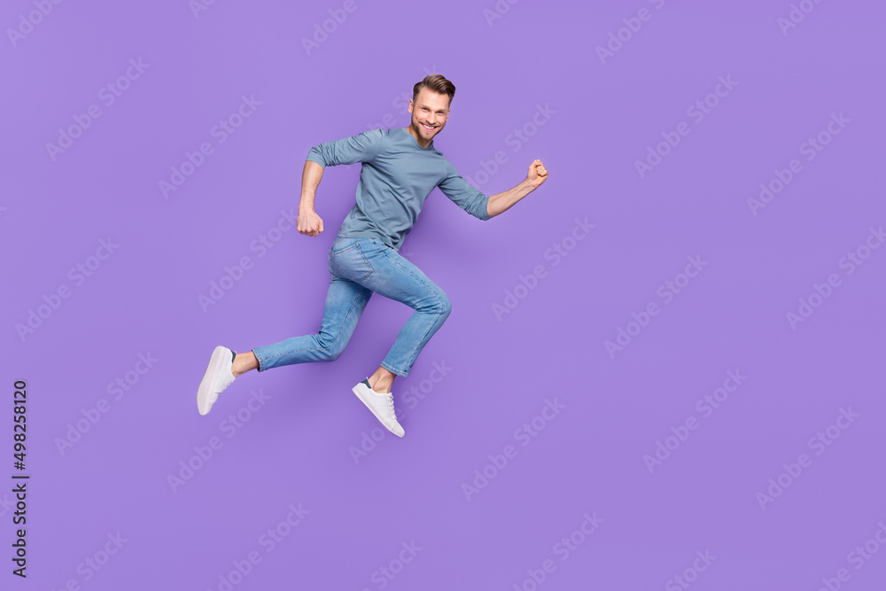 Photo of excited attractive guy wear grey shirt running fast jumping high empty space isolated violet color background Photo of excited attractive guy wear grey shirt running fast jumping high empty space isolated violet color background
