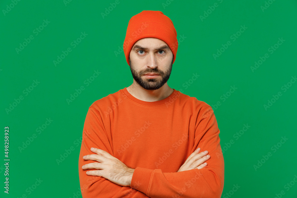 Young strict sad offended serious caucasian man 20s wearing orange sweatshirt hat hold hands crossed folded look camera isolated on plain green background studio portrait. People lifestyle concept Young strict sad offended serious caucasian man 20s wearing orange sweatshirt hat hold hands crossed folded look camera isolated on plain green background studio portrait. People lifestyle concept