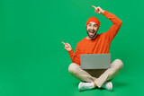 Full body young surprised amazed happy man 20s in orange sweatshirt hat sit hold use work on laptop pc computer poin index finger aside on workspace isolated on plain green background studio portrait