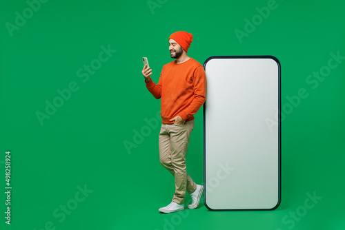 Full body young smiling man 20s wear orange sweatshirt hat stand near big blank screen mobile cell phone with workspace copy space mockup area use smartphone isolated on plain green background studio. photo