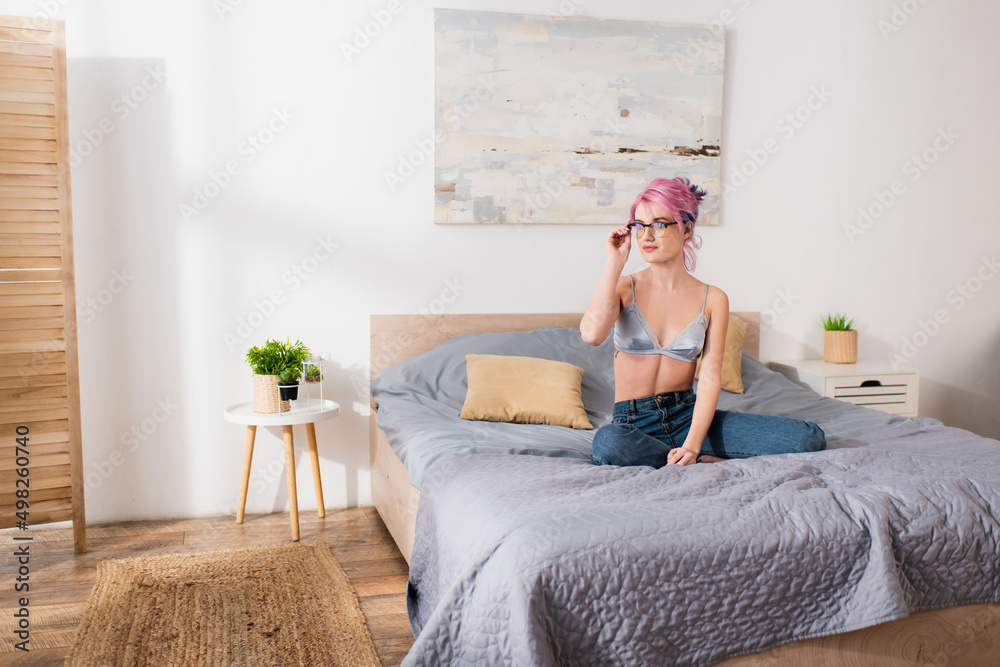 young woman with pink hair adjusting eyeglasses and sitting on bed.
