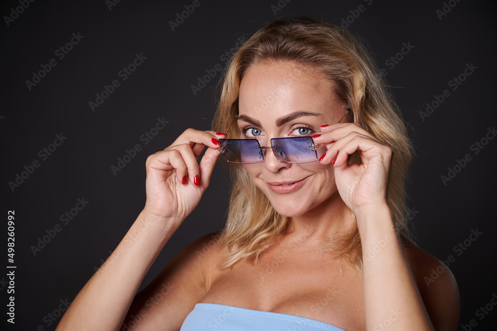 Headshot beautiful blonde European woman confidently looking at camera through blue sunglasses, isolated over black background with copy space for ads Headshot beautiful blonde European woman confidently looking at camera through blue sunglasses, isolated over black background with copy space for ads