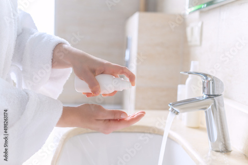 Woman in the robe washing hands using liquid soap or antibacterial gel for protective and care dry skin near white sink in bathroom with sunlight from the window.