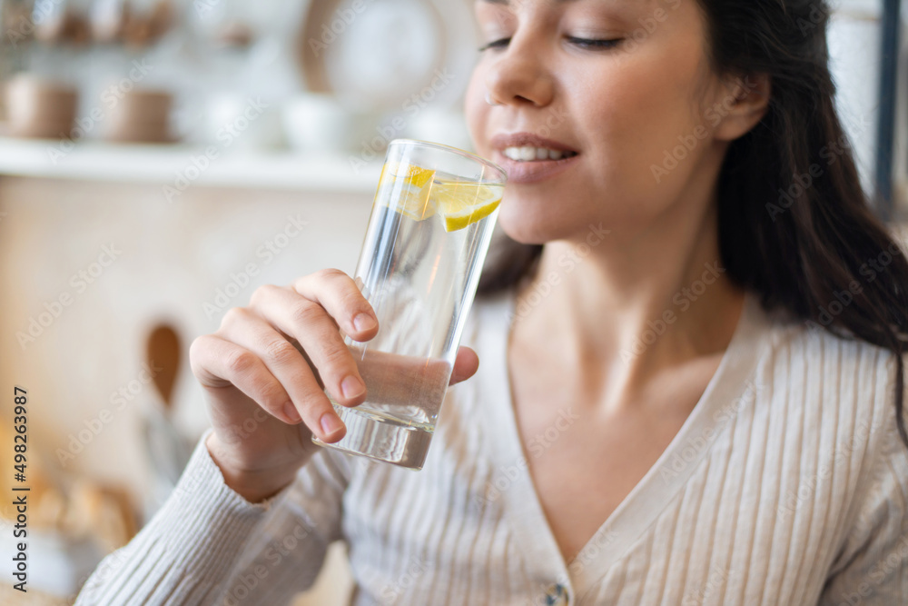 Refreshment concept. Young woman drinking water with lemon, holding glass, standing in kitchen, closeup Refreshment concept. Young woman drinking water with lemon, holding glass, standing in kitchen, closeup