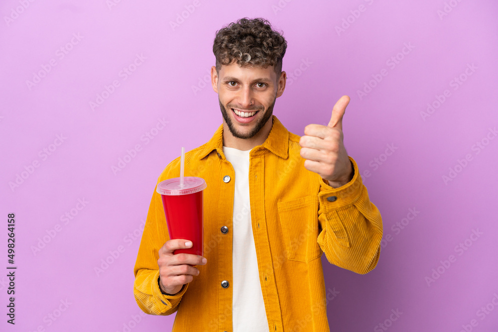Young blonde man holding soda isolated on purple background with thumbs up because something good has happened Young blonde man holding soda isolated on purple background with thumbs up because something good has happened