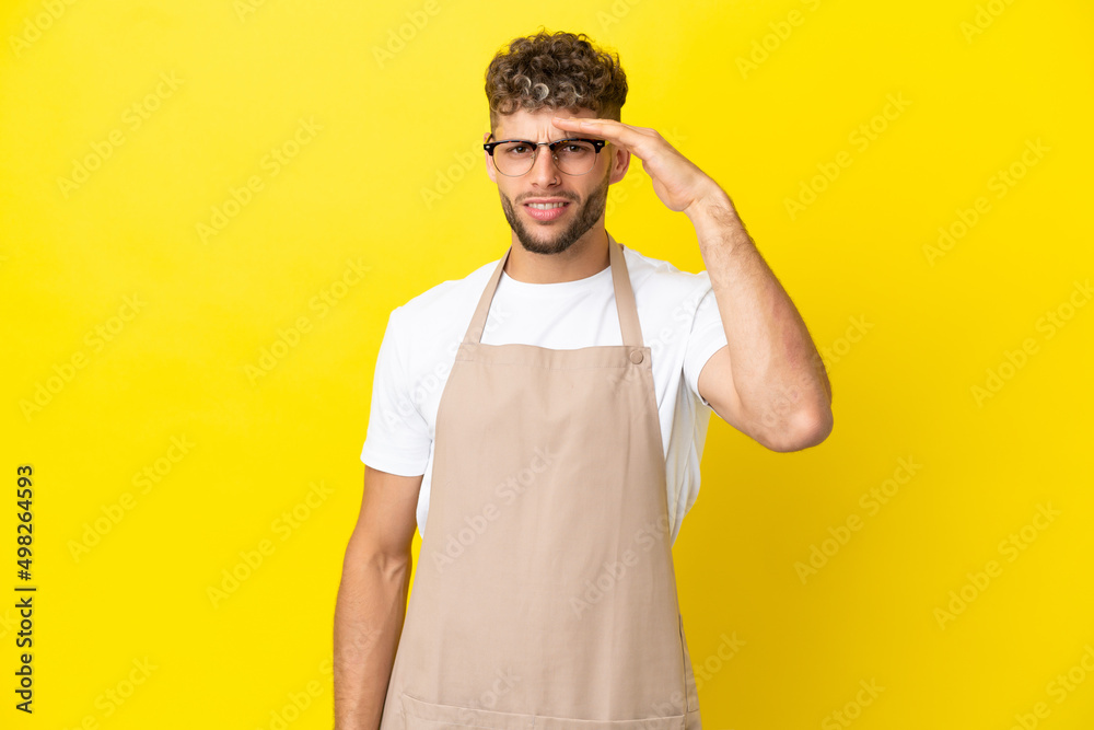 Restaurant waiter blonde man isolated on yellow background looking far away with hand to look something Restaurant waiter blonde man isolated on yellow background looking far away with hand to look something