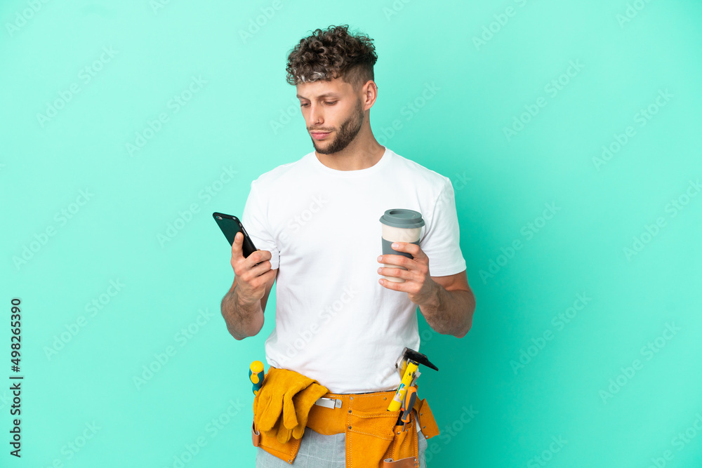 Young electrician blonde man isolated on green background holding coffee to take away and a mobile Young electrician blonde man isolated on green background holding coffee to take away and a mobile