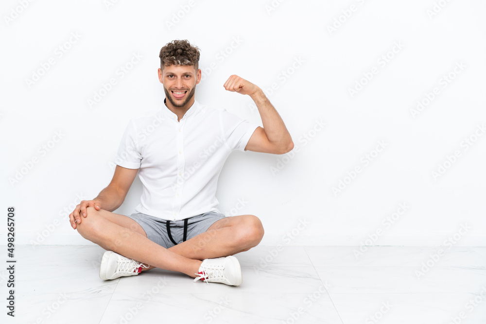 Young blonde man sitting on the floor isolated on white background doing strong gesture Young blonde man sitting on the floor isolated on white background doing strong gesture