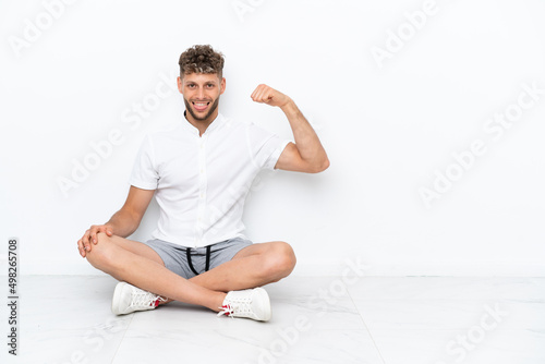 Young blonde man sitting on the floor isolated on white background doing strong gesture