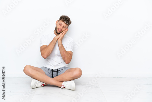 Young blonde man sitting on the floor isolated on white background making sleep gesture in dorable expression
