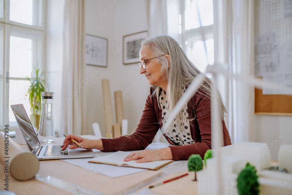 Senior woman architect with model of houses working on laptop in office.